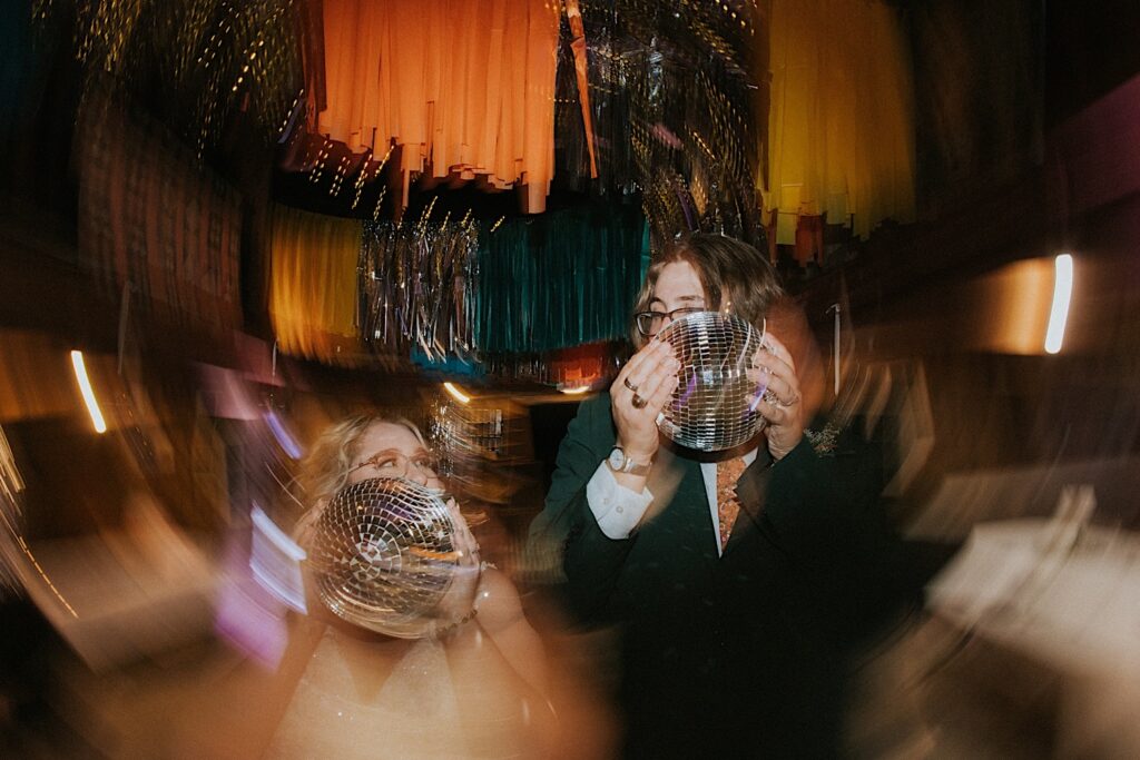 A bride and groom hold disco balls in front of their faces and look at one another while light distorts around them during their indoor wedding reception at the Clayville Historic Site