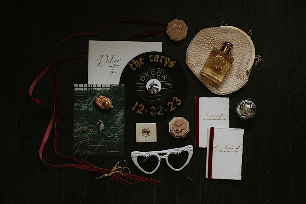A wedding flatlay consisting of sunglasses, wedding invites, perfume, a vinyl record, rings, and more