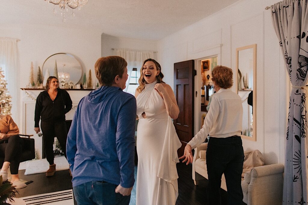A bride laughs while standing in a living room in her wedding dress as friends and family see her for the first time