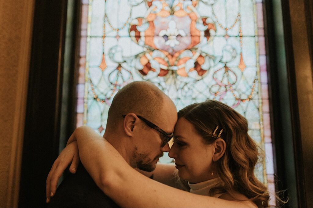 A bride and groom touch foreheads together while standing in front of a stained glass window inside of the Vrooman Mansion