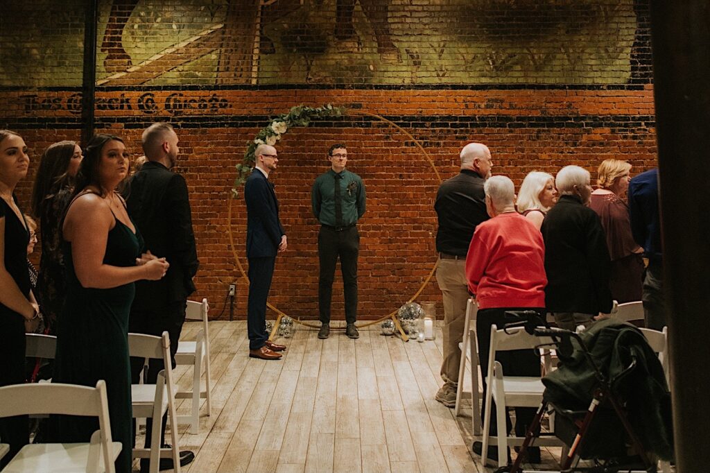 A groom and officiant stand at the indoor ceremony space at Reality on Monroe as guests watch during an intimate wedding ceremony