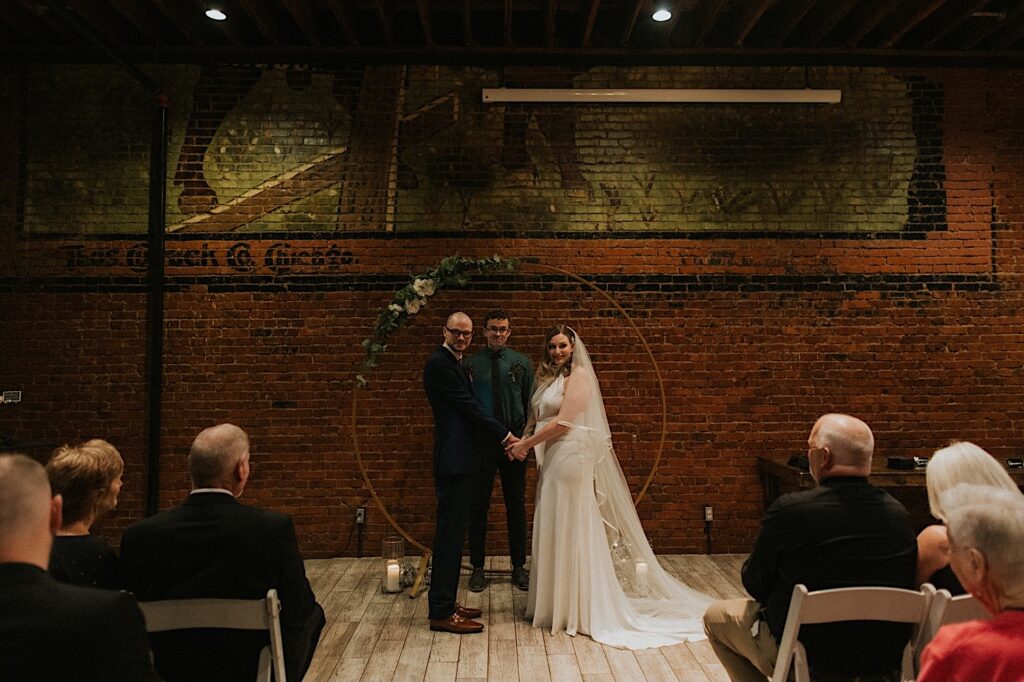 A bride and groom hold hands and look at their guests during their intimate wedding ceremony at Reality on Monroe