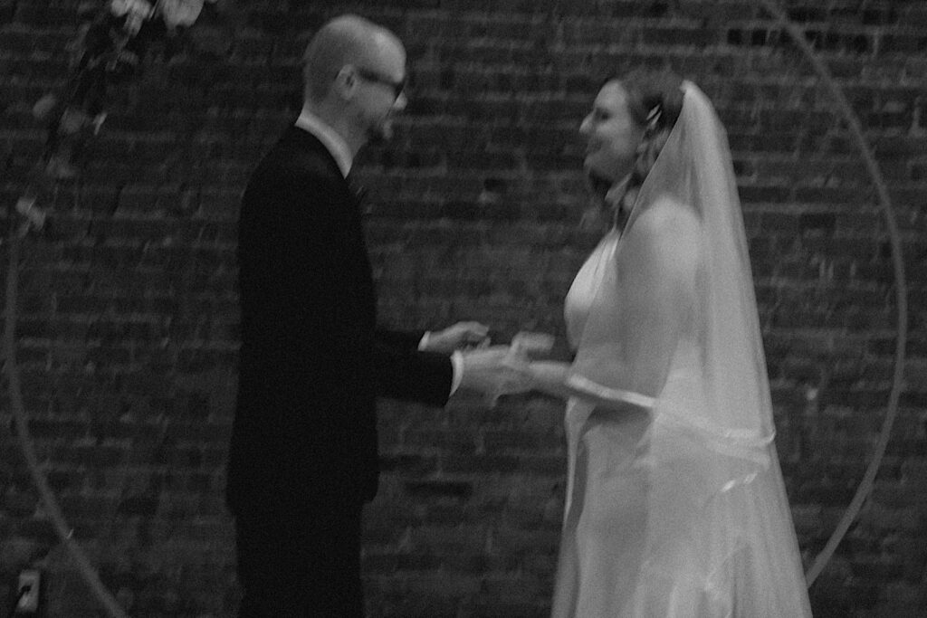 Black and white photo of a bride and groom smiling at one another and holding hands during their intimate indoor wedding ceremony at Reality on Monroe