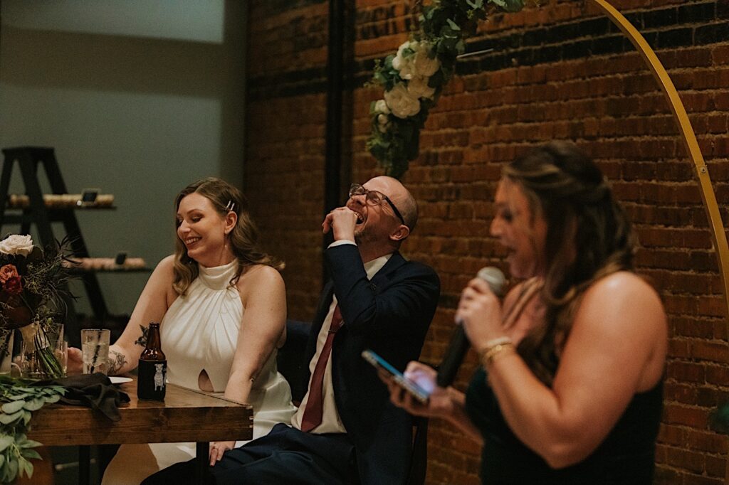 A bride and groom sitting at their sweetheart table laugh as a woman gives a speech during their intimate wedding reception at Reality on Monroe