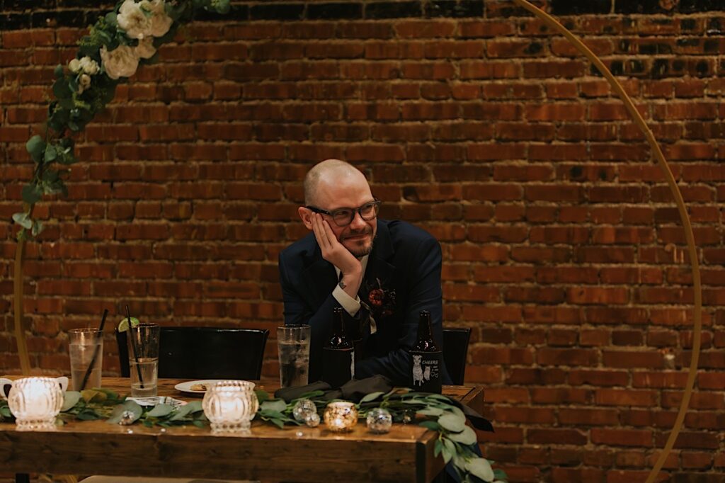 A groom smiles as he sits at the sweetheart table and watches the bride dance during their intimate wedding reception at Reality on Monroe
