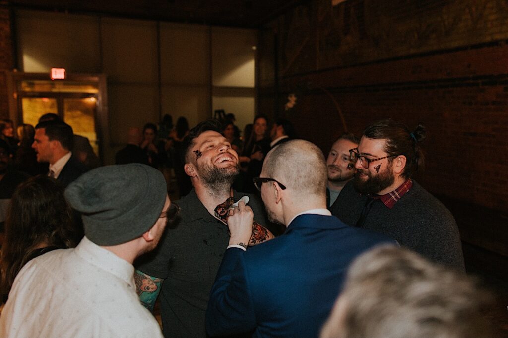 A man smiles as the groom helps apply a temporary tattoo on his neck as other guests watch during an intimate wedding reception at Reality on Monroe
