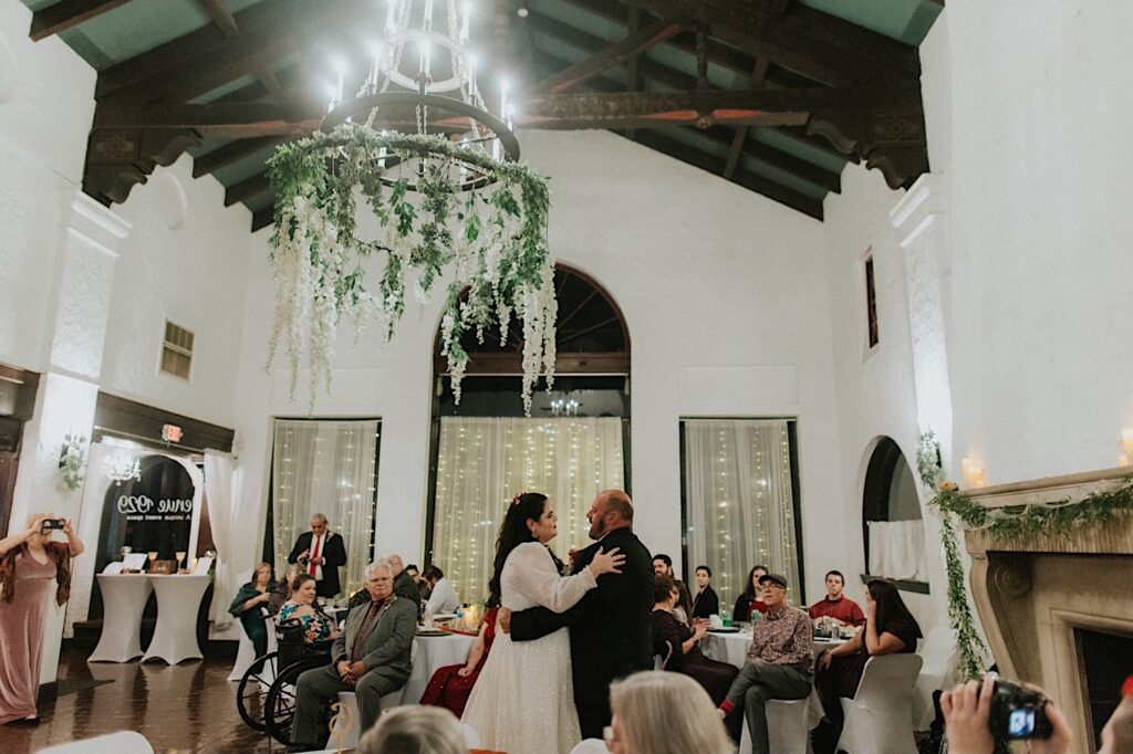 A bride dances with her father during an indoor wedding reception as guests sit and watch around them