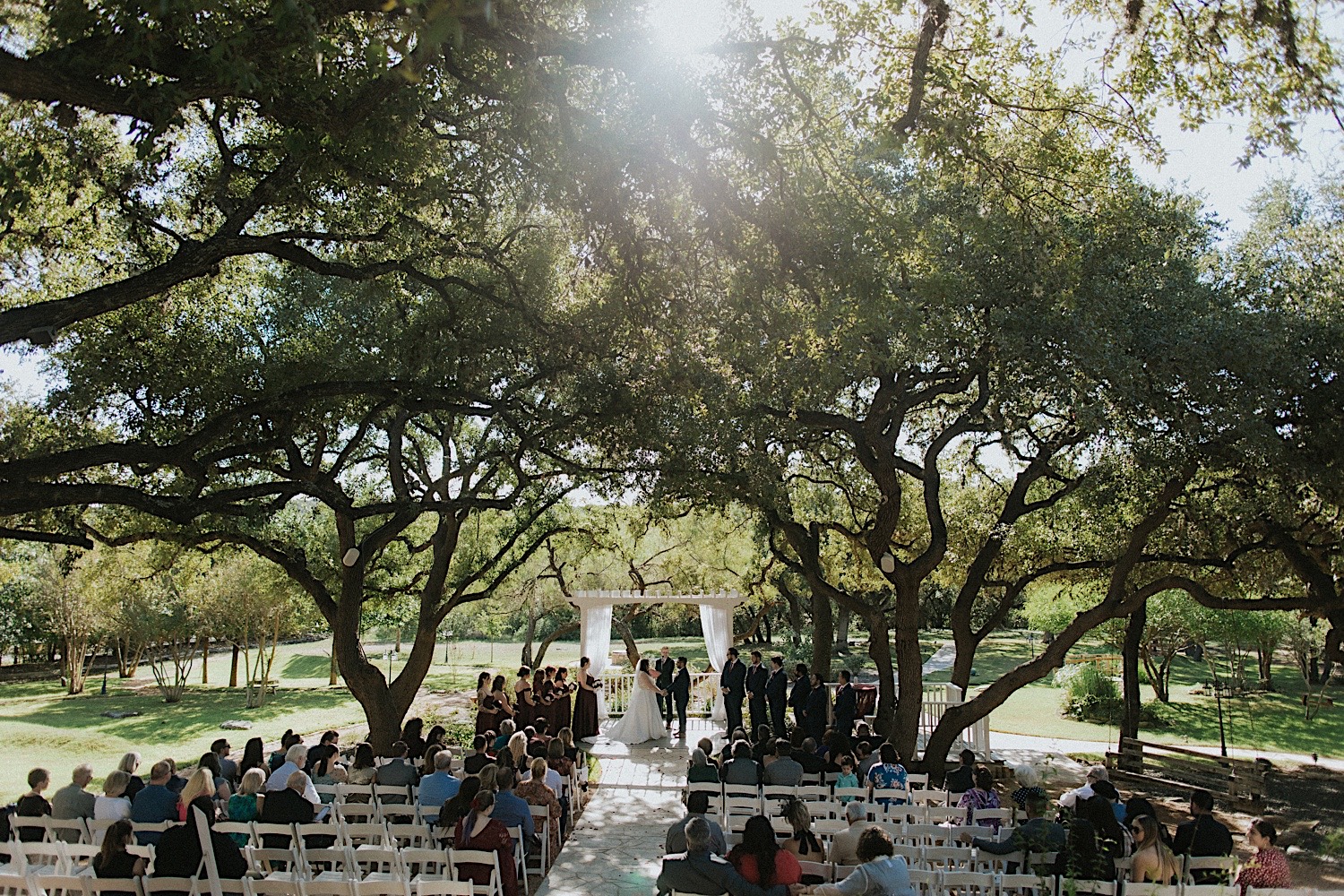 A wedding ceremony takes place outdoors underneath the cover of large trees in a park