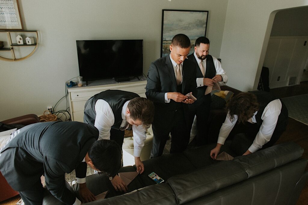 A groom stands with his 4 groomsmen as they all fold their pocket kerchiefs together in the living room