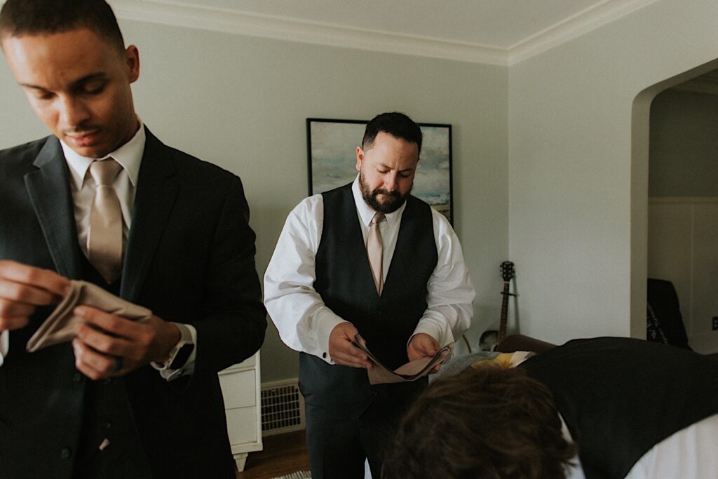A groom folds his pocket kerchief in a living room alongside some of his groomsmen