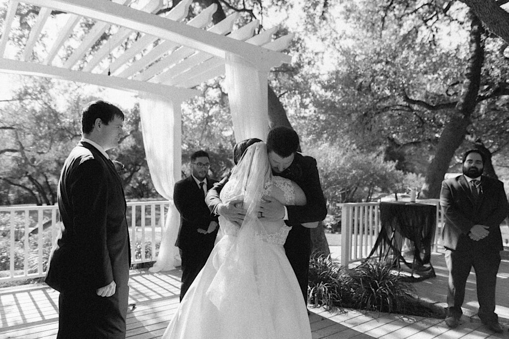 Black and white photo of a bride and groom hugging one another during their outdoor wedding ceremony