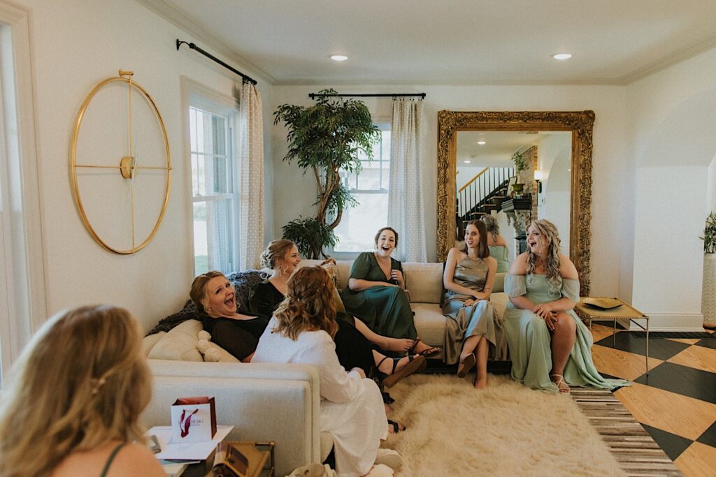 A bride sits with her 6 bridesmaids on a couch as they all laugh together