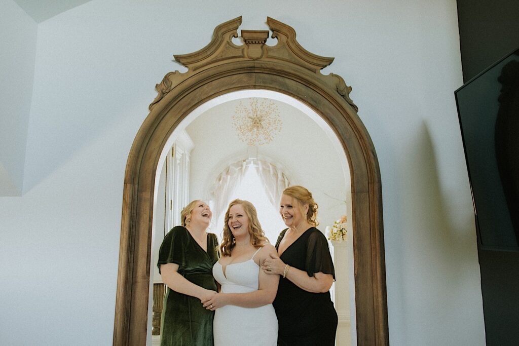 A bride laughs while standing in the mirror in her wedding dress alongside her mother and sister who are also laughing