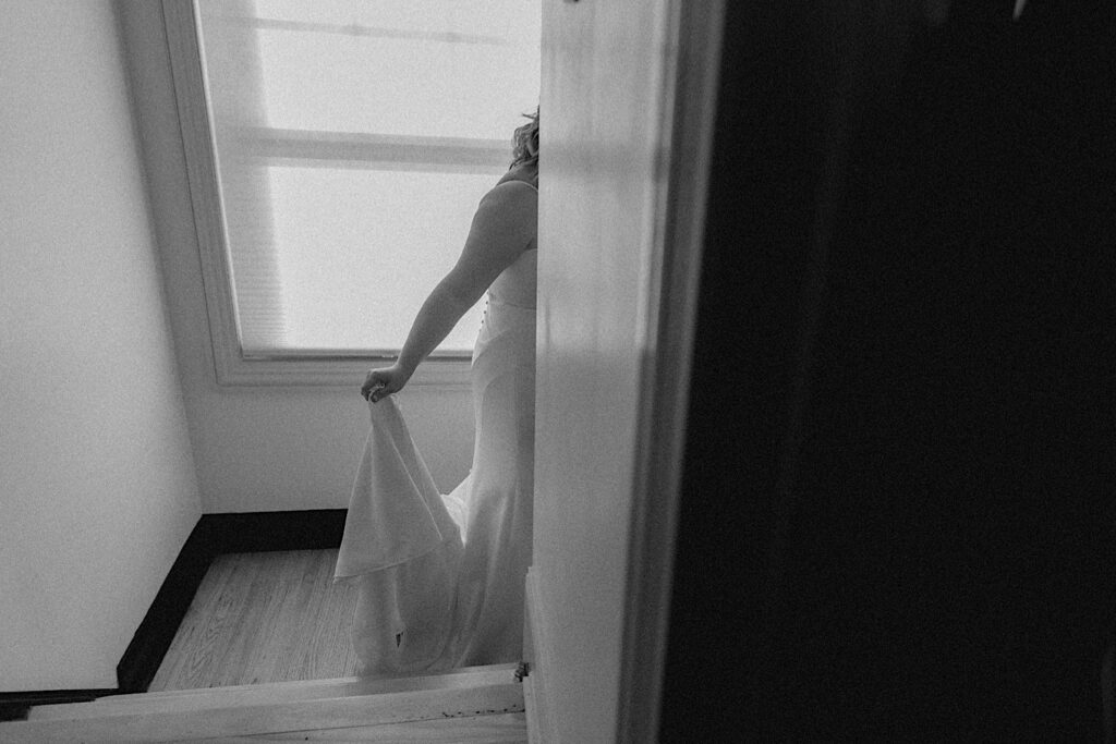 Black and white photo of a bride holding the back of her wedding dress as she walks down the stairs