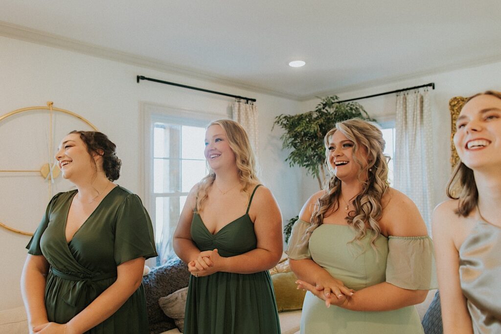 4 bridesmaids stand side by side in a living room and smile as they see a bride in her wedding dress for the first time