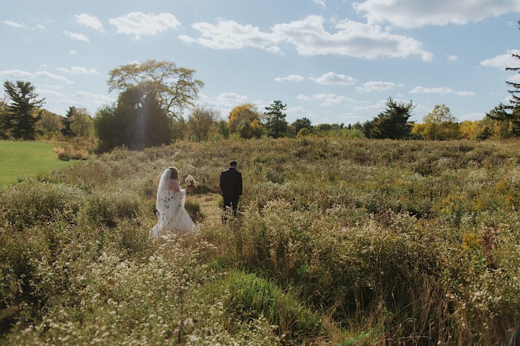 A bride walks up behind the groom as he stands in a field on a sunny day before their first look