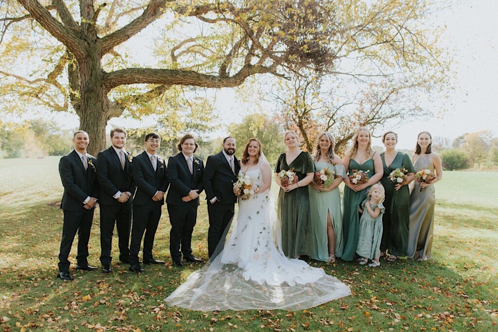 A bride and groom stand with their wedding parties on either side of them in a park as they all smile at the camera
