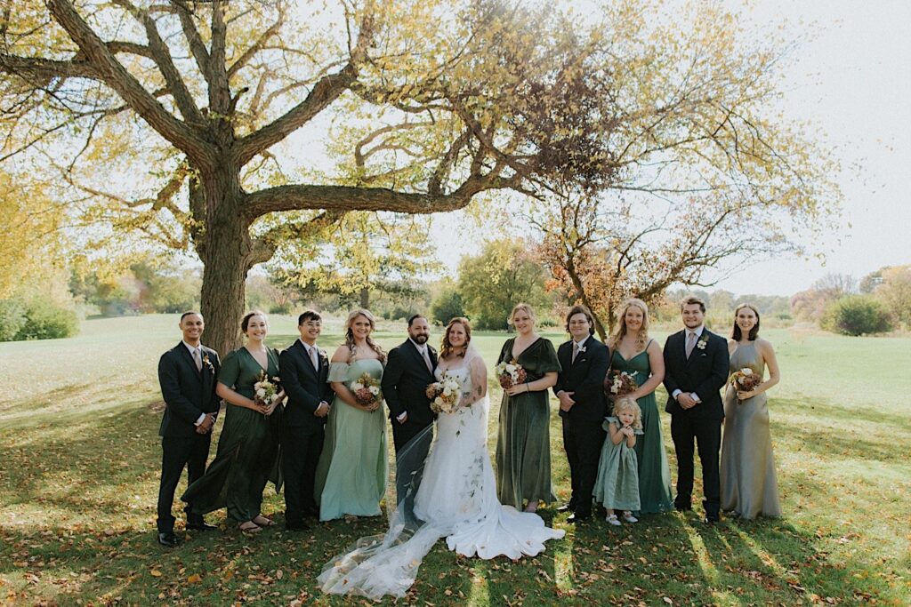 A bride and groom stand with their wedding parties on either side of them in a park as they all smile at the camera