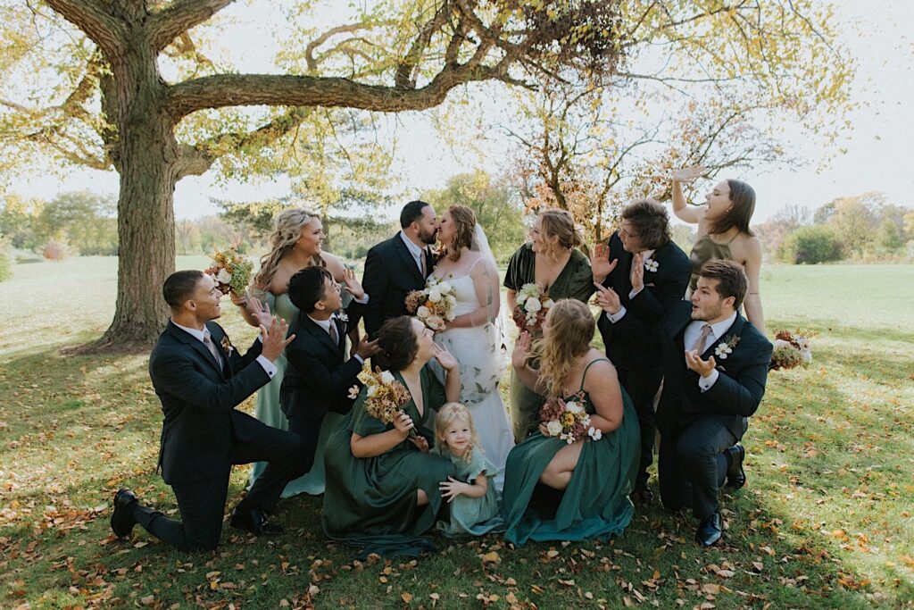 A bride and groom kiss as members of their wedding party surround them and cheer while all standing in a park together