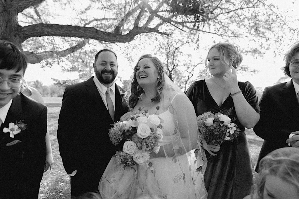 Black and white photo of a bride and groom laughing while members of their wedding party smile around them 