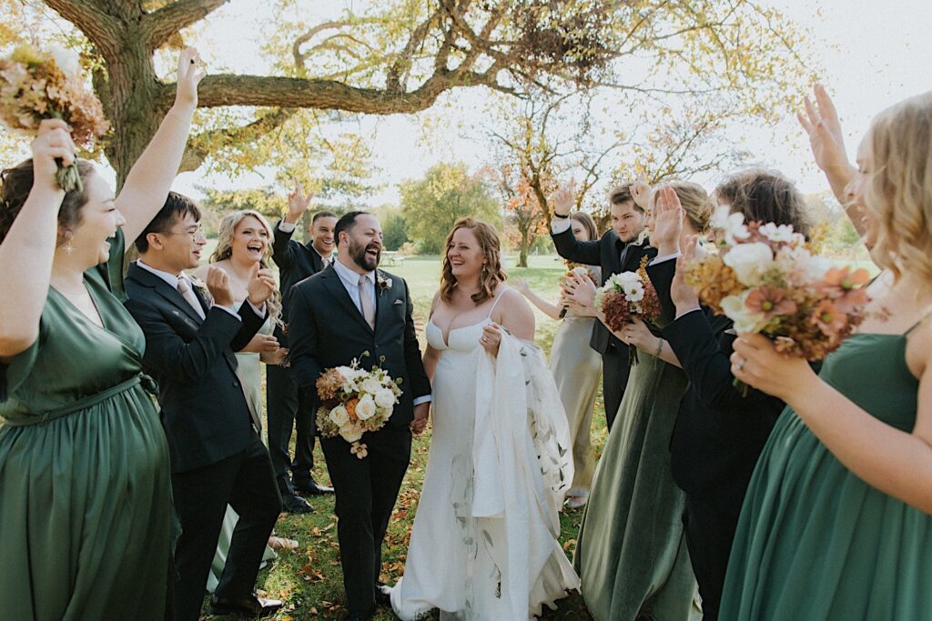 A bride and groom laugh while standing in between members of their wedding parties who all cheer for them while inside of a park
