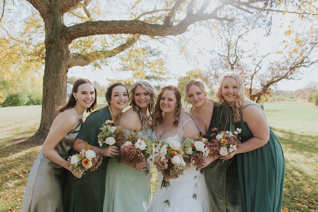 A bride and her 5 bridesmaids smile at the camera while standing in a park together