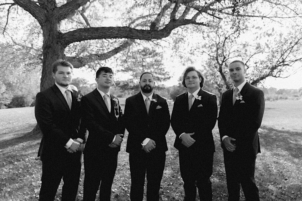 A black and white photo of a groom and his 4 groomsmen standing in a park and posing for a portrait photo