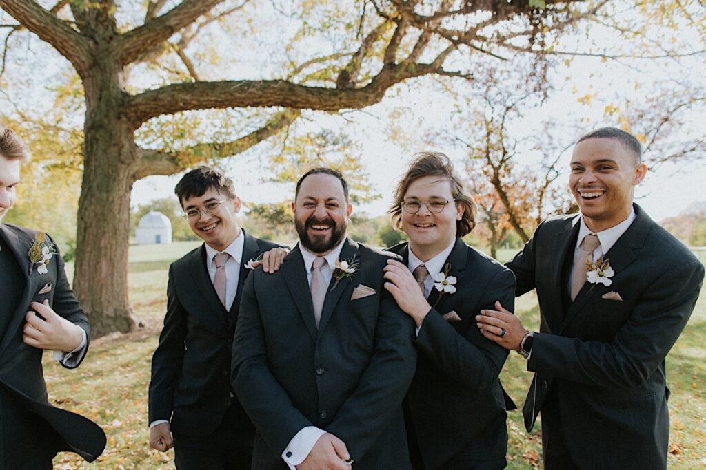 A groom smiles while his 4 groomsmen put their hands on his shoulders while taking portraits in a park