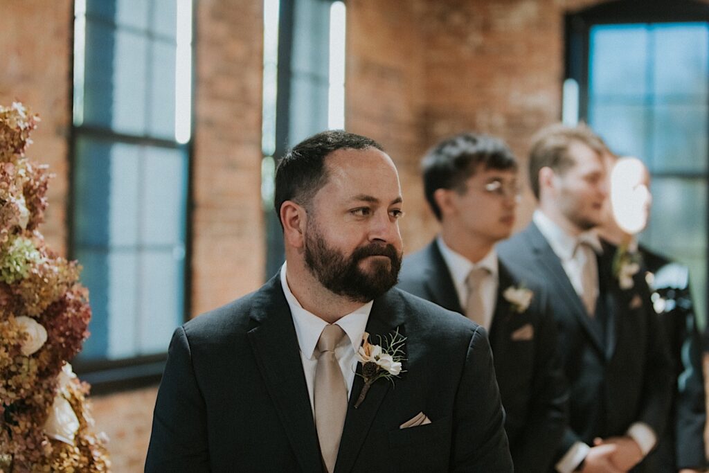 A groom smiles with his groomsmen next to him as the bride enters for their wedding ceremony inside of Trailside Event Center
