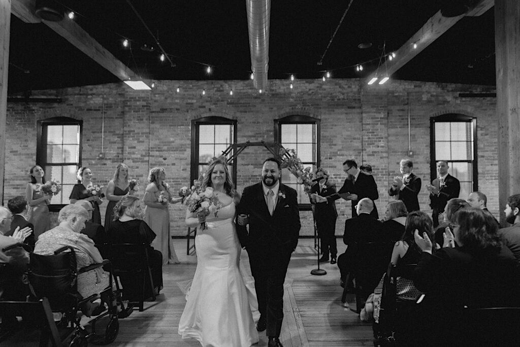 Black and white photo of a bride and groom walking down the aisle together after their wedding ceremony inside of Trailside Event Center