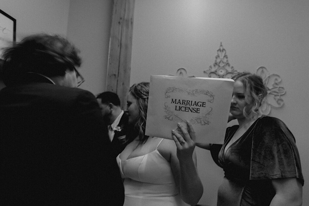 Black and white photo of a bride holding her marriage license while inside Trailside Event Center with members of her wedding party as well as the groom next to her
