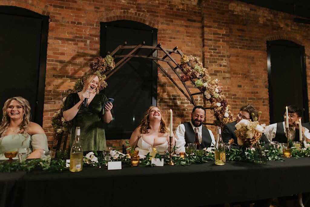 A bride and groom laugh while sitting at the head table with their wedding party as a bridesmaid gives a speech during a wedding reception at Trailside Event Center