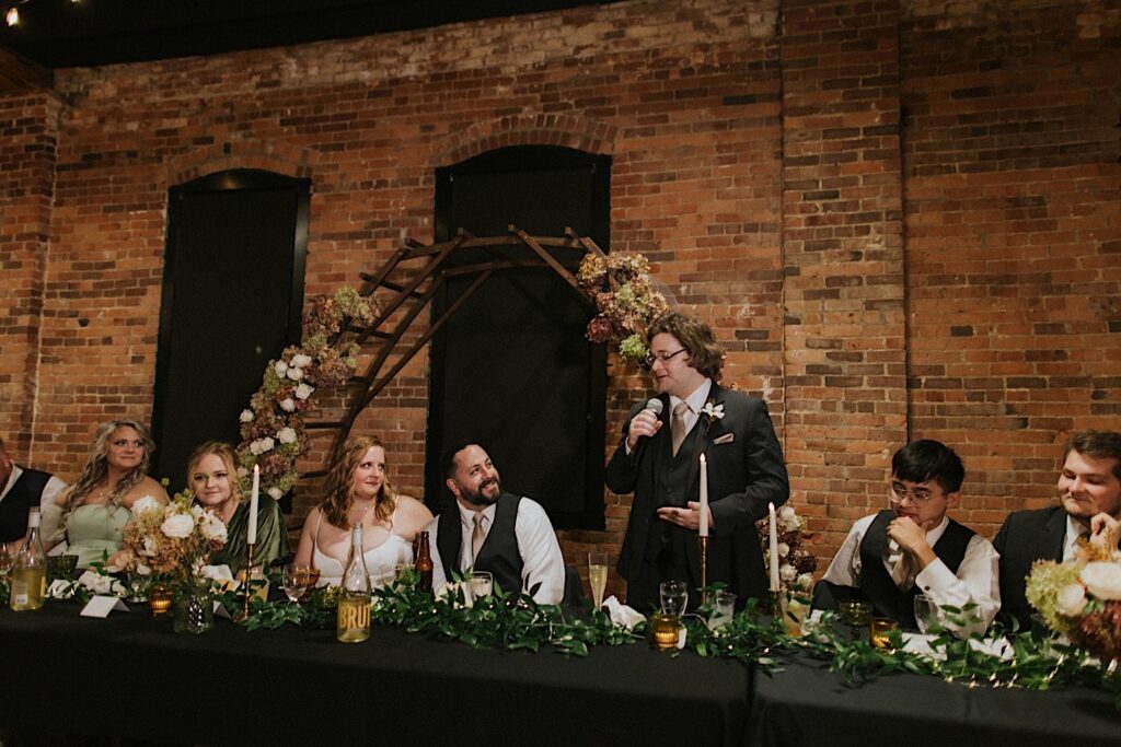 A bride and groom sit at their head table with their wedding party members as a groomsmen gives a speech during their reception at Trailside Event Center