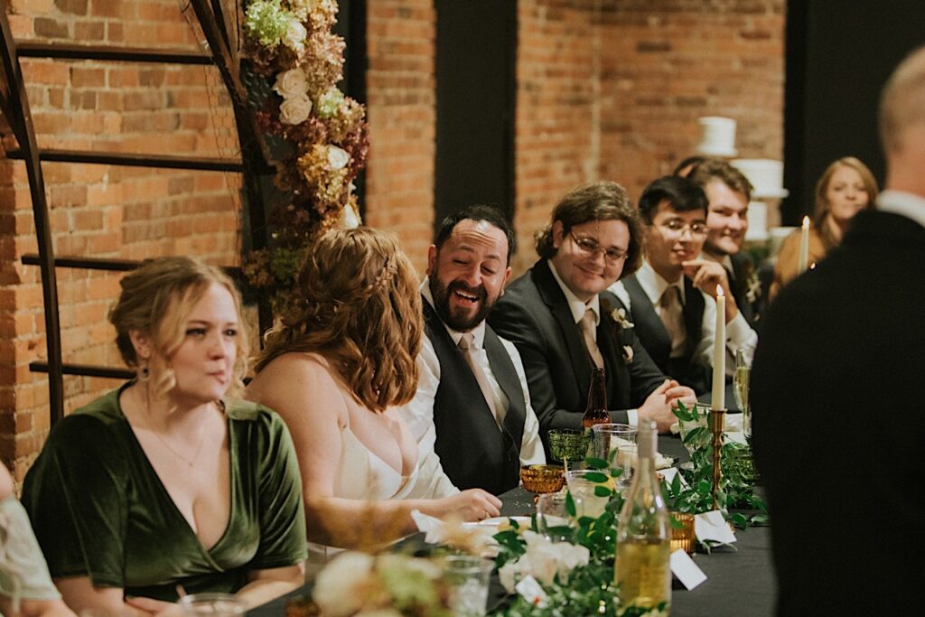 A bride and groom laugh at one another while sitting at their head table with members of their wedding parties as a man gives a speech during a wedding reception at Trailside Event Center
