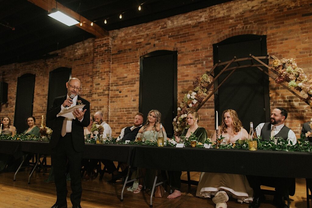 A father gives a speech during a wedding reception at Trailside Event Center as the bride and groom and wedding party members listen