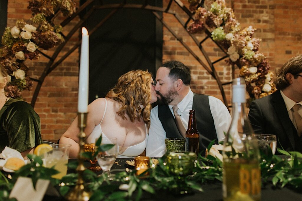 A bride and groom kiss one another while sitting at the head table of their wedding reception at Trailside Event Center