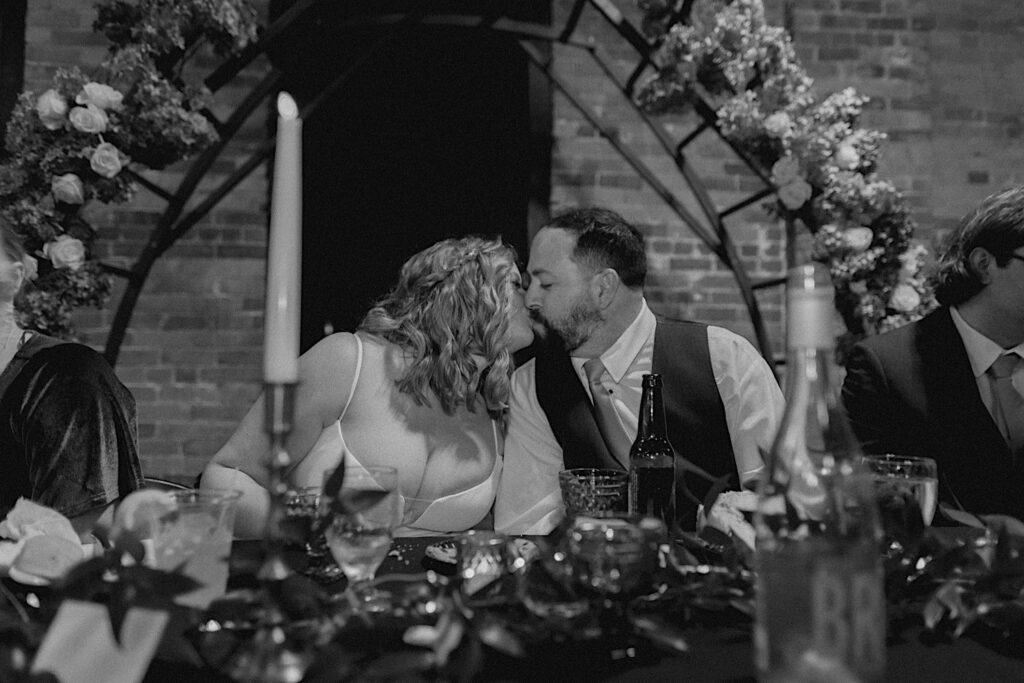 Black and white photo of a bride and groom kiss one another while sitting at the head table of their wedding reception at Trailside Event Center