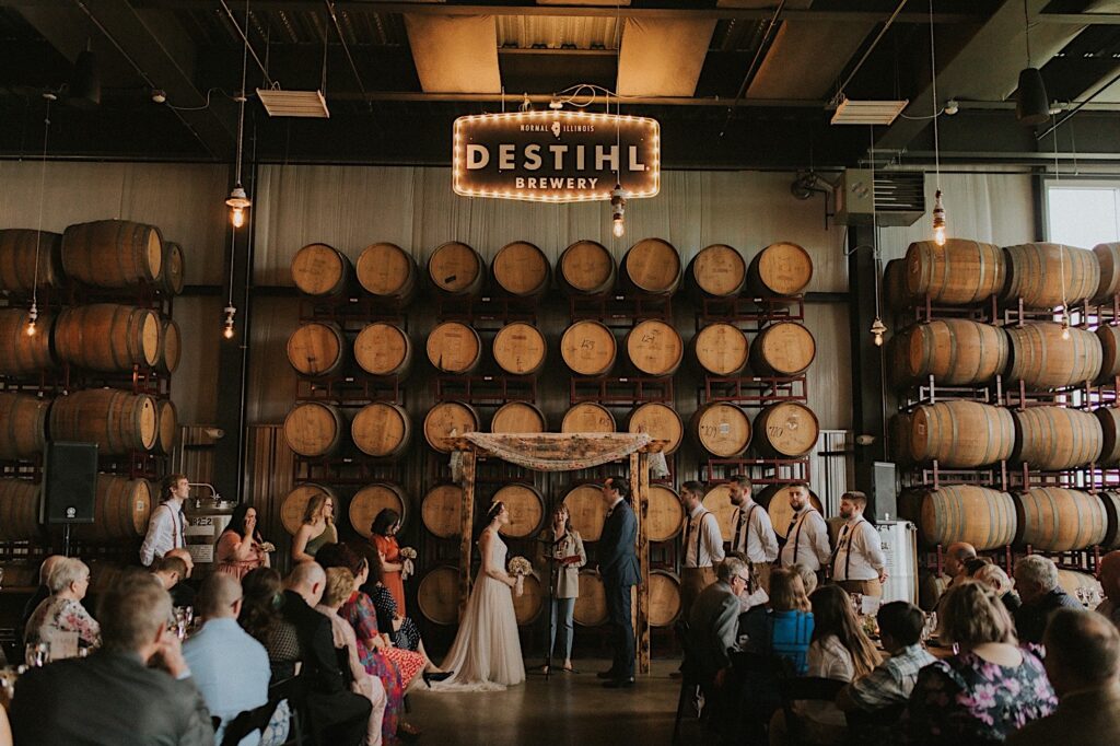 A wedding ceremony takes place inside of The Barrel Room at the wedding venue, The Destihl Brewery in Central Illinois