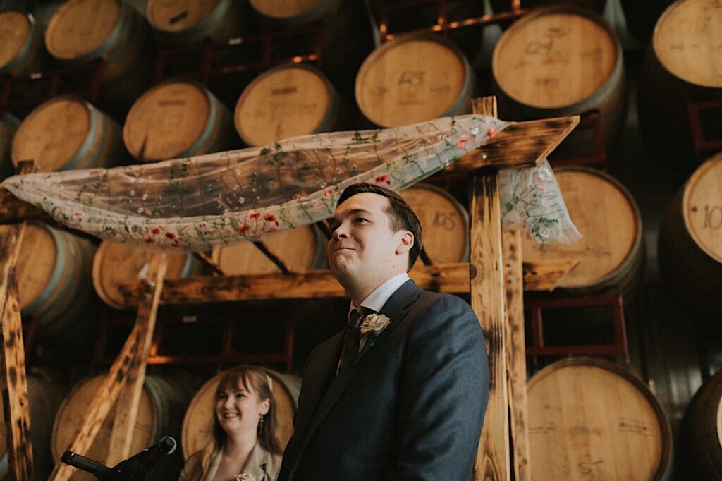 A groom smiles while looking towards the bride during their ceremony inside of their wedding venue, The Barrel Room in the Destihl Brewery of Central Illinois