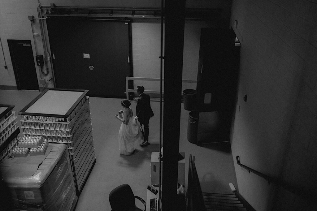 Black and white photo of a bride and groom walking through the beer packaging area of their wedding venue, the Destihl Brewery in Central Illinois