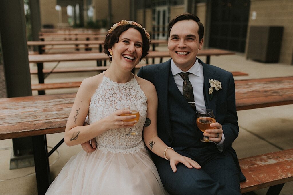 A bride and groom sit on a picnic table and smile at the camera while holding glasses of beer outside of their wedding venue, the Destihl Brewery in Central Illinois