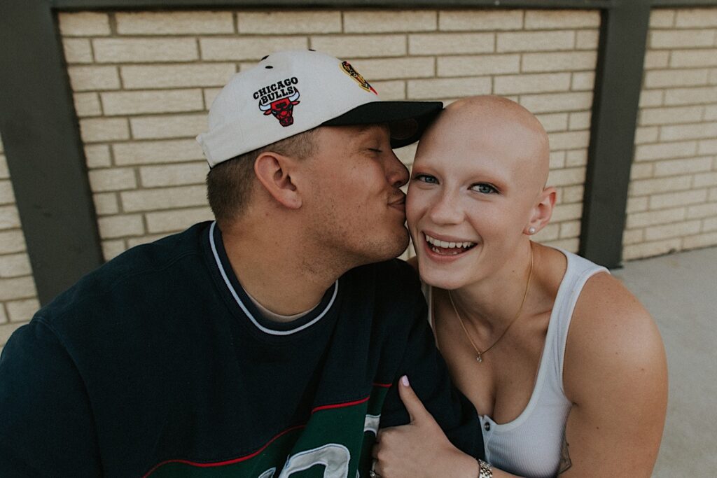 A woman smiles as a man kisses her cheek as the two sit on a curb outside of The Rootbeer Stand in Central Illinois during their engagement session