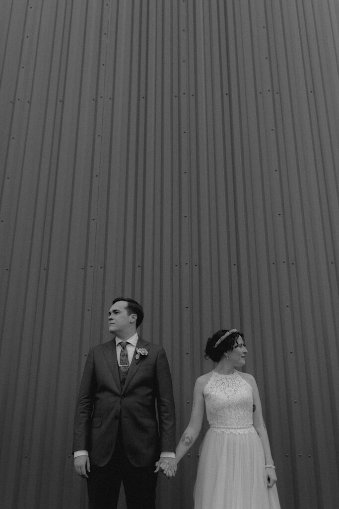 Black and white photo of a bride and groom holding hands while outside of a metal building