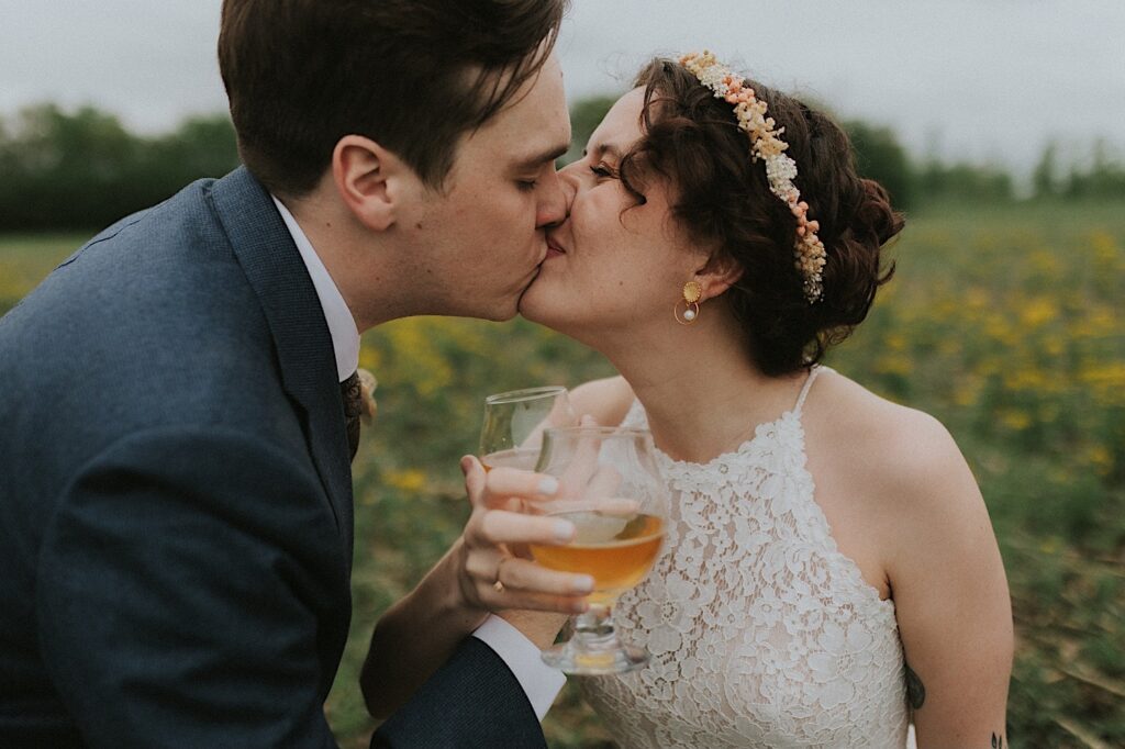 A bride and groom kiss one another while outside of their wedding venue, the Destihl Brewery of Central Illinois, in their hands are glasses of beer