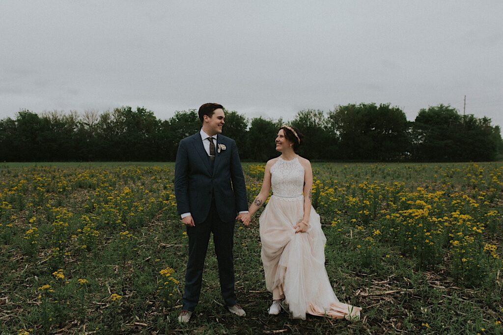 A bride and groom hold hands and smile while standing in a field of yellow flowers outside of their wedding venue, the Destihl Brewery in Central Illinois
