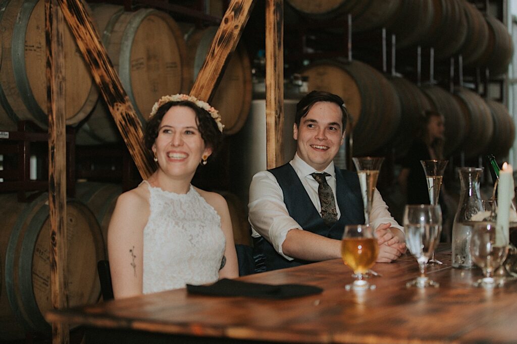 A bride and groom smile as they sit next to one another at a table for their reception at their wedding venue, The Barrel Room of the Destihl Brewery
