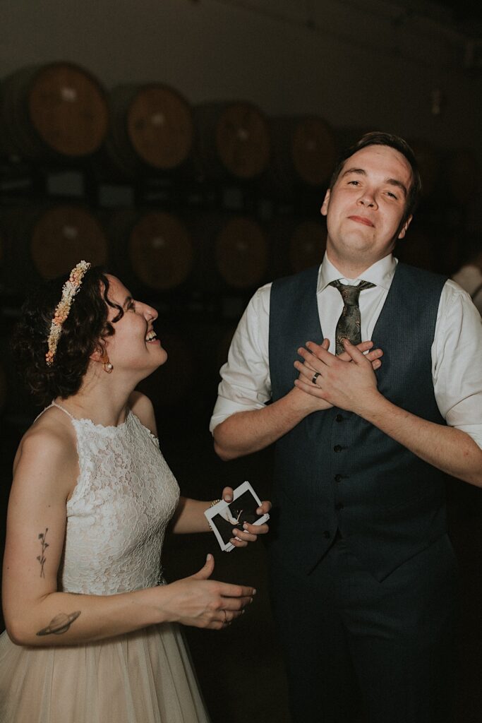 A bride smiles at the groom while he holds his heart and looks at the camera after seeing polaroid photos that the photographer took of the couple throughout their wedding day