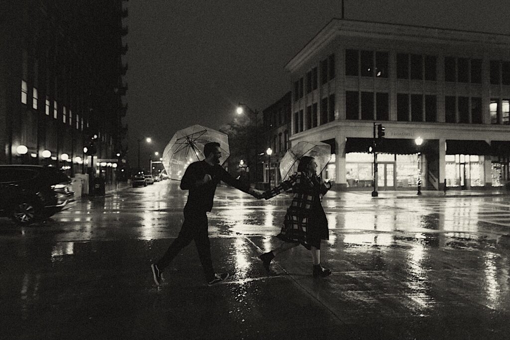 A couple runs during a rainy evening across the street with clear umbrellas in their hands.