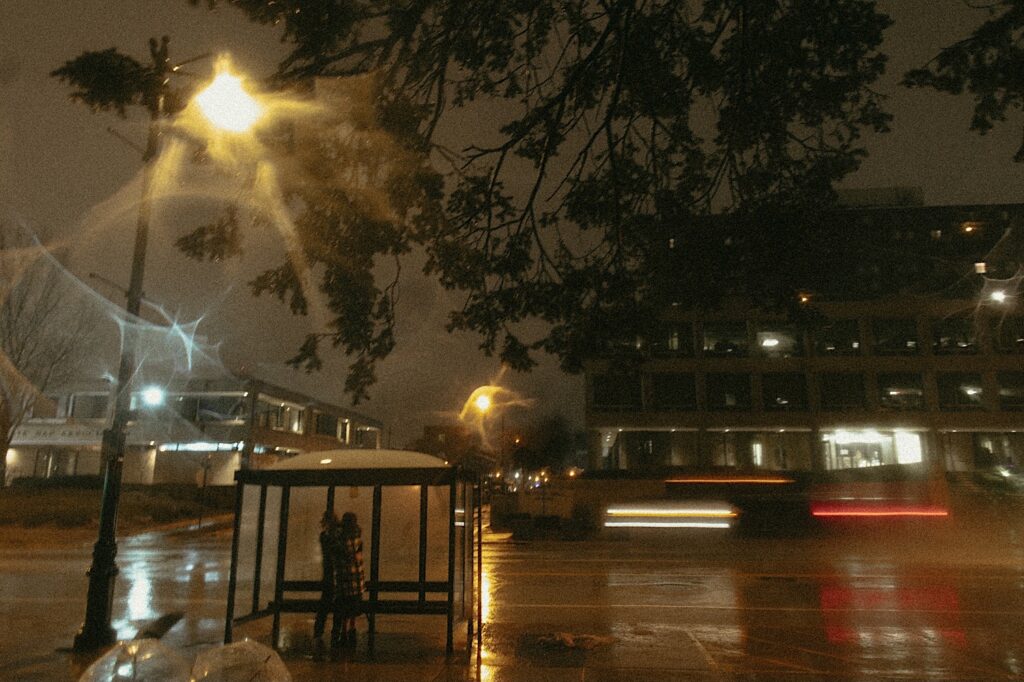 Couple stands under a bus stop with cars whizzing by and lights flashing during a couples session in downtown Springfield in the rain. 