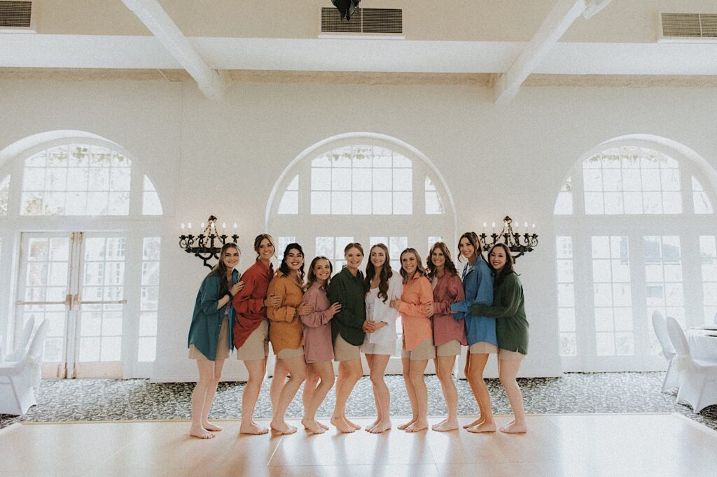 Bride stands surrounded by her bridesmaids dressed in different colored oversized button ups inside beautiful white room with large windows. 